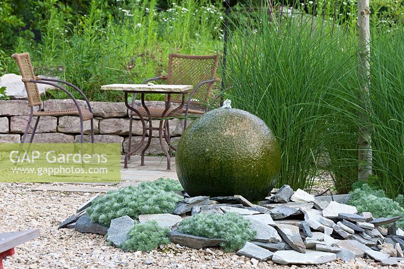 Wasserspiel aus Granitkugel und Schindeln. Gartenmöbel aus Metall mit Bistrotisch neben Trockenmauer. Artemisia schmidtiana 'Nana' und Miscanthus sinensis