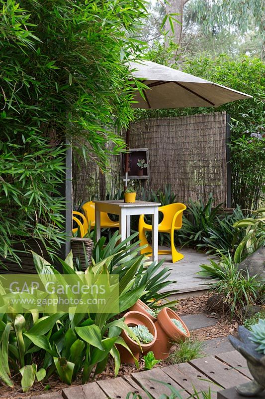 Holz Holzterrasse bedeckt von einem großen weißen Regenschirm mit leuchtend gelben Plastik-Retro-Stühlen und einem Holztisch, abgeschirmt mit Bambus-Streichholzjalousien und eingerahmt von einem Klumpen aus Bambus und Aspidistra elatior, Gusseisenpflanze.