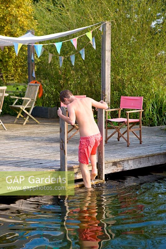 Der Junge verlässt das 'natürliche' Schwimmbad, indem er mit einem Sonnensegel auf eine Leiter klettert, die an einem Sitzbereich mit Holzdecks aus Holz befestigt ist