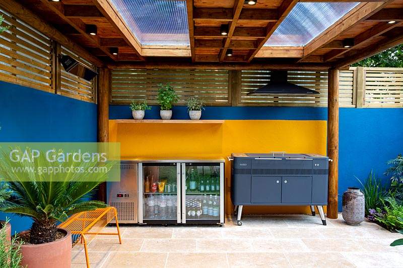 Außenraum mit Grill und Kühlschrank. Umgeben von einem modernen Holzgitterzaun und einer blau gestrichenen Wand.