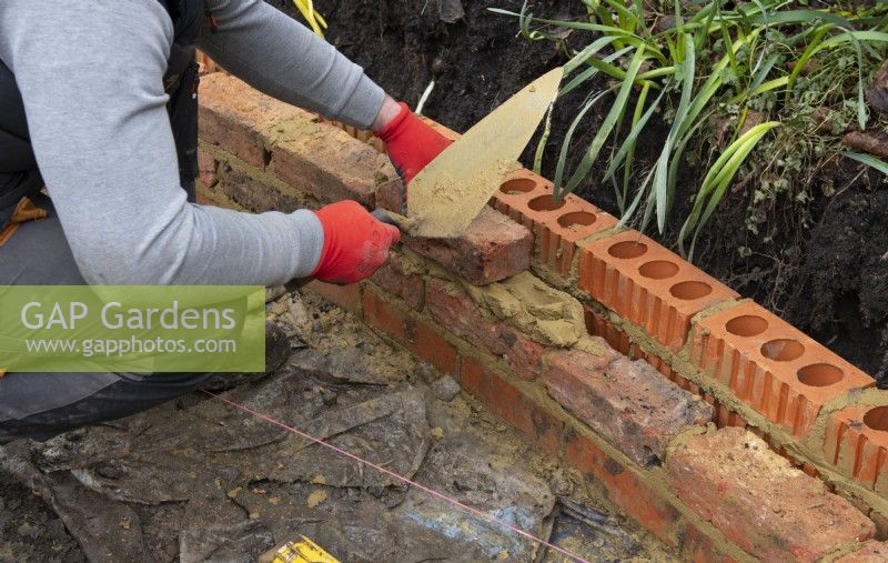 Ein Arbeiter, der während der Renovierung eines kleinen Londoner Gartens mit einer Maurerkelle eine niedrige Ziegelmauer um ein Hochbeet baut.