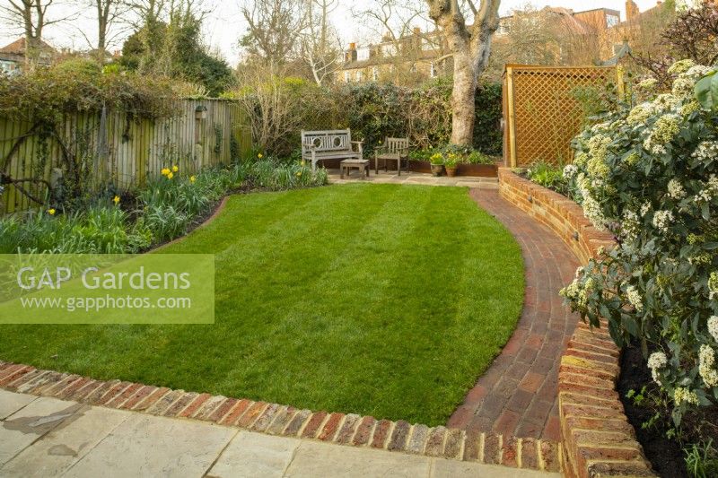 Ein 'Nachher-Foto' eines kleinen Londoner Gartens nach einer Renovierung, die eine neue niedrige Backsteinmauer, einen geschwungenen Pfad, eine erweiterte Terrasse und einen neu verlegten Rasen umfasste.