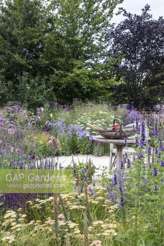 Staudenbeete mit Salvia, Achillea, Agastache und Verbena bonariensis wickeln sich um einen Holztisch mit einem Korb voller Gartengeräte. Carol Klein, RHS-Ikone des Gartenbaus – RHS Hampton Court Palace Garden Festival 2023 