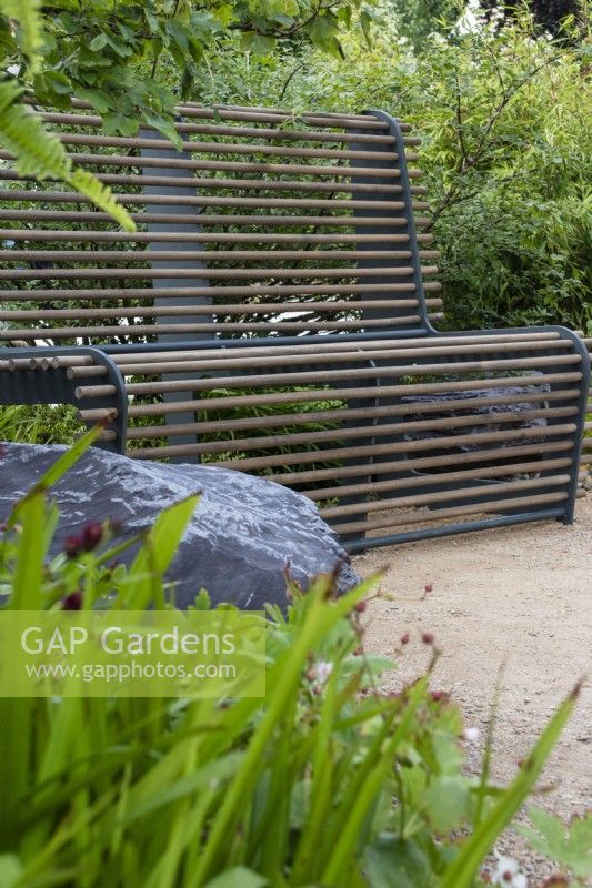 Ein Sitz mit Metallrahmen und Holzstäben inmitten von Pflanzen – Cancer Research UK Legacy Garden – Designer Paul Hervey-Brookes – RHS Hampton Court Flower Palace Garden Festival 2023. 
