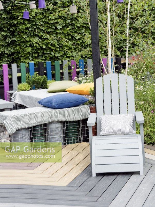 Eine Terrasse aus verschiedenfarbigen Holzdielen, die in einem Muster verlegt sind, mit Gabionenbänken und Kissen darauf, Sommer Juli 