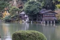 Blick über den Kasumigaike-Teich auf das Teehausgebäude namens Uchihashitei und auf mehrere Bäume mit Wigwams aus Bambus und Seilen zum Schutz vor Schneeschäden namens Yukitsuri. 