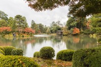 Blick über den Kasumigaike-Teich auf die Insel Horaijima und das Gebäude am Teichufer namens Uchihashitei. Bäume mit Herbstfärbung. 